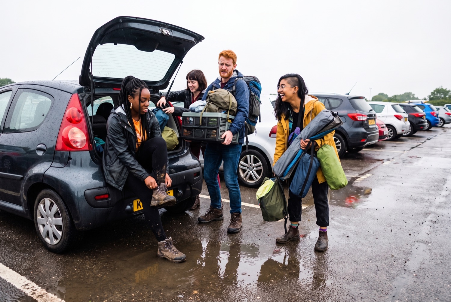 Four friends unloading rucksacks, a folded tent and a crate from a small hatchback in a wet car park.
