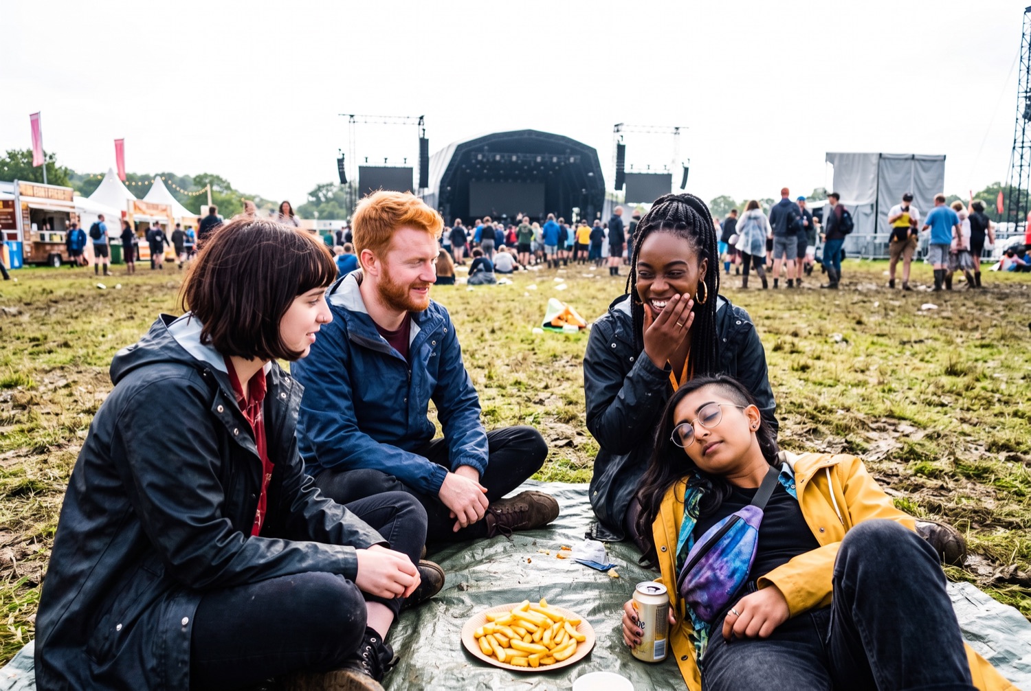 Four friends sitting on a groundsheet on trampled grass between sets, a paper plate of chips between them, a distant stage behind.