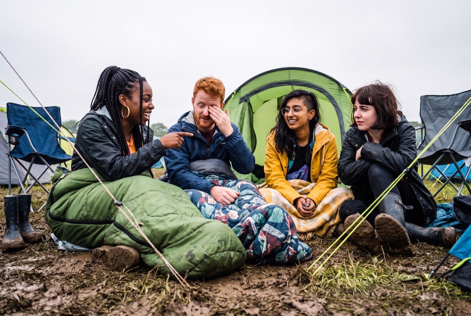 Four friends sitting on the ground outside a tent in flat morning light, wrapped in sleeping bags, one of them laughing at another mid-story.